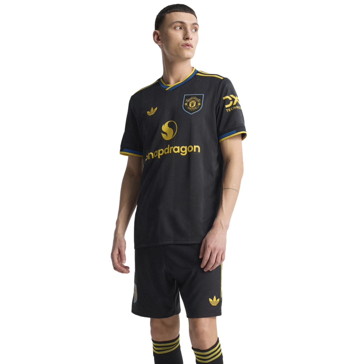 A man models the black Manchester United 2024/25 third kit, featuring yellow and blue trim, the Adidas Trefoil logo, and Snapdragon sponsor.