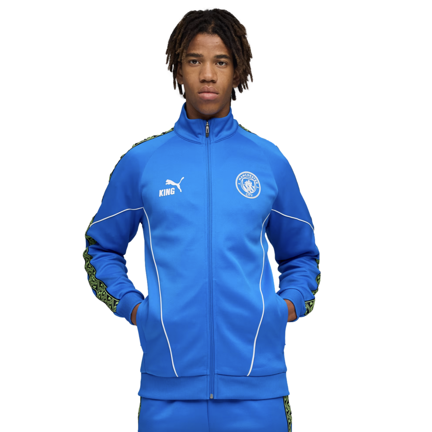 Young man with dreadlocks wearing a blue Manchester City tracksuit featuring a Puma logo and club crest, standing against a black background.
