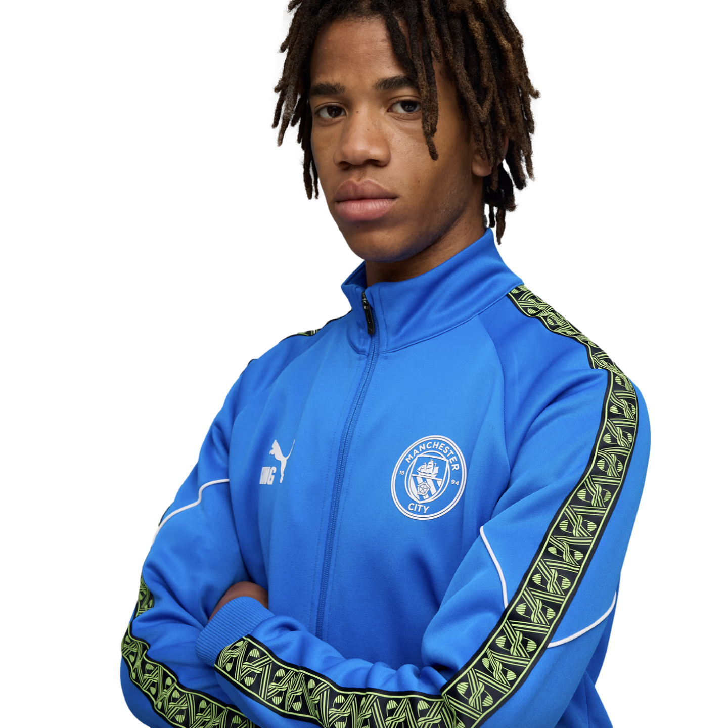 Young man with dreadlocks wearing a blue Manchester City tracksuit jacket, posing with arms crossed against a black background.