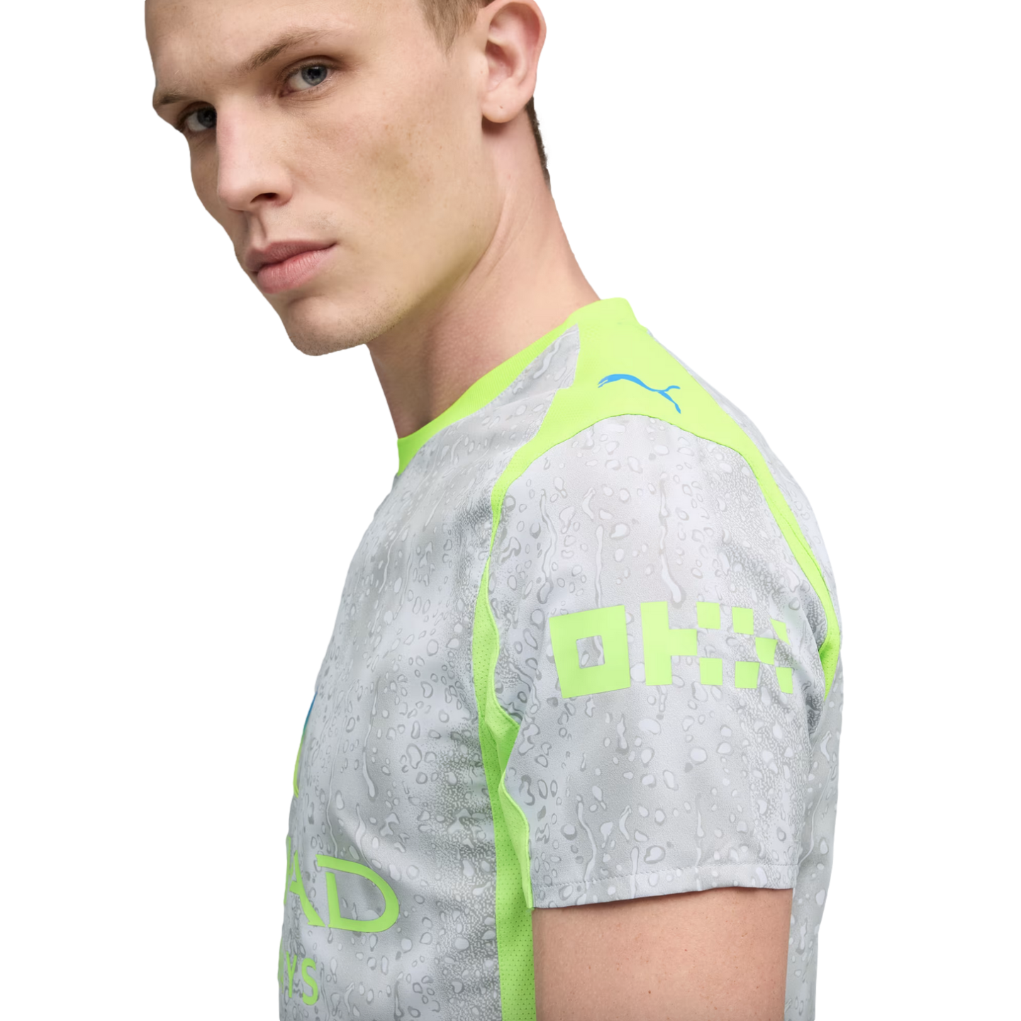 A fair-skinned man with blond hair wears a grey and lime green Puma sports jersey featuring a water droplet pattern and sponsorship branding.