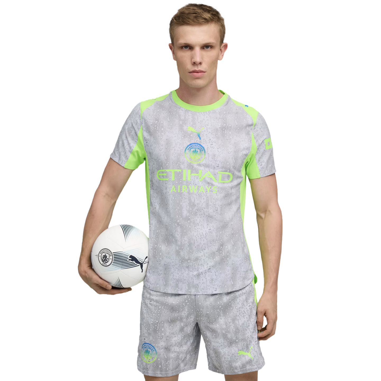 Man in a Manchester City soccer kit, grey with neon green accents, holding a soccer ball under his arm against a black background.