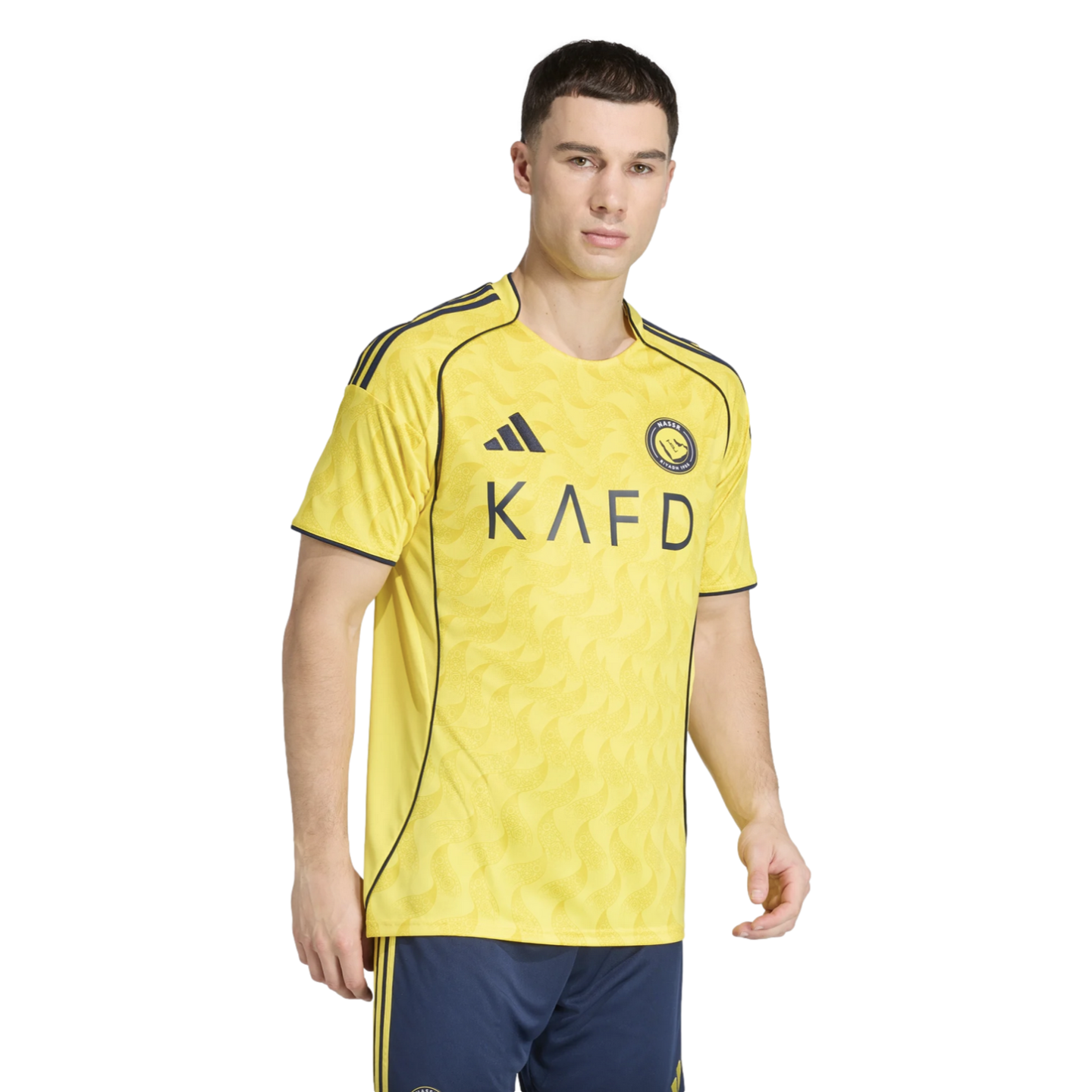 A man modeling a yellow soccer jersey with navy blue trim and "KAFD" across the chest, paired with navy blue shorts. The jersey features a subtle wavy pattern.