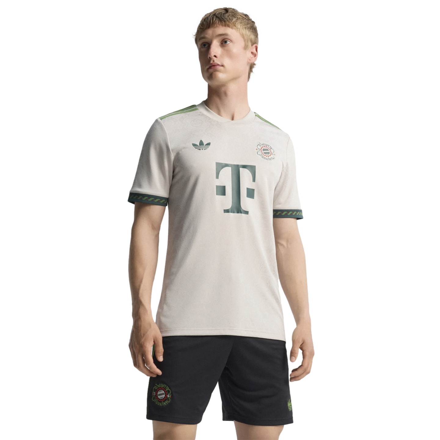 A man models a cream-colored Bayern Munich soccer jersey with green accents and a central "T" logo, paired with black shorts against a solid black background.