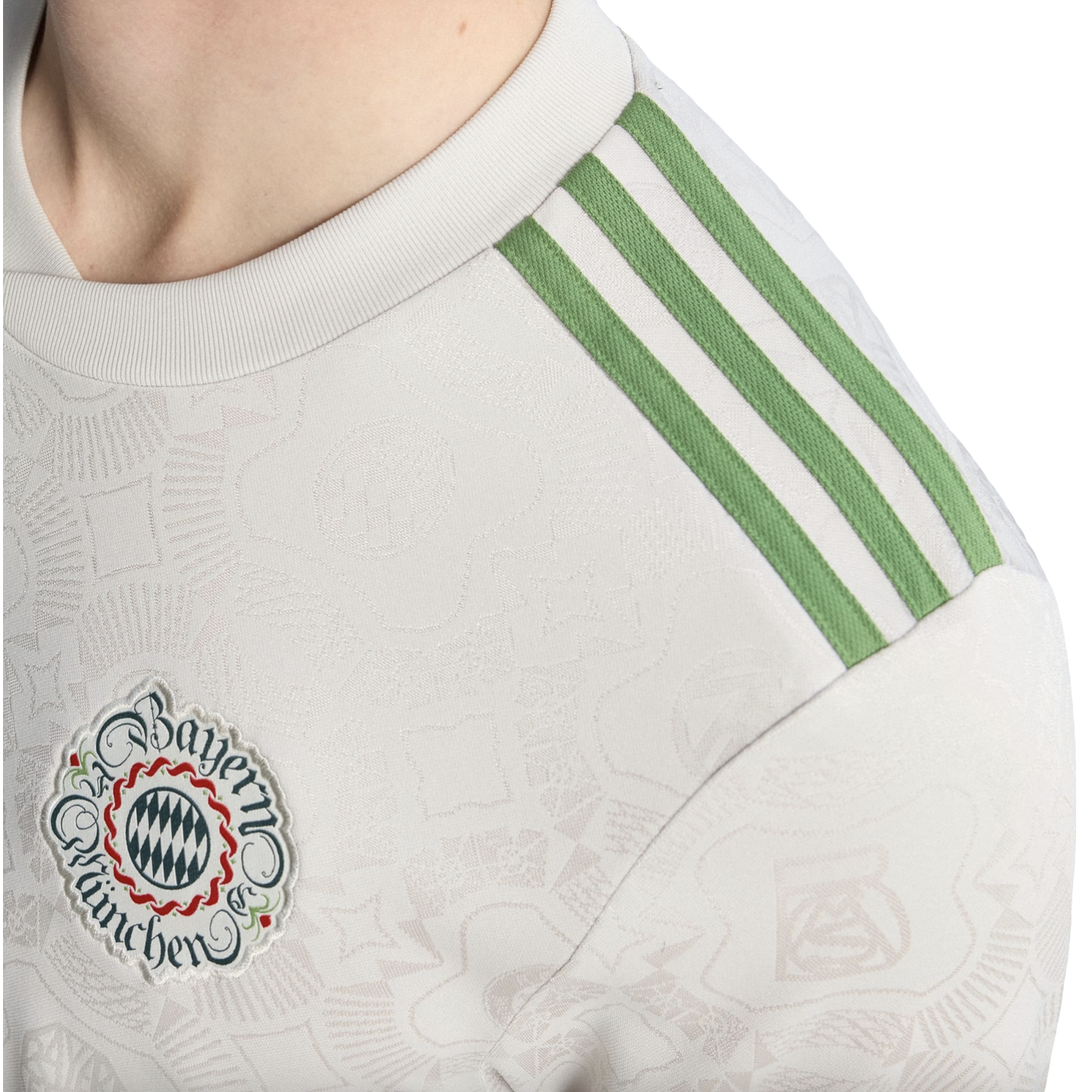 Close-up of a white Bayern Munich jersey featuring green Adidas shoulder stripes and an intricate club crest patch. The fabric has a subtle, embossed pattern.
