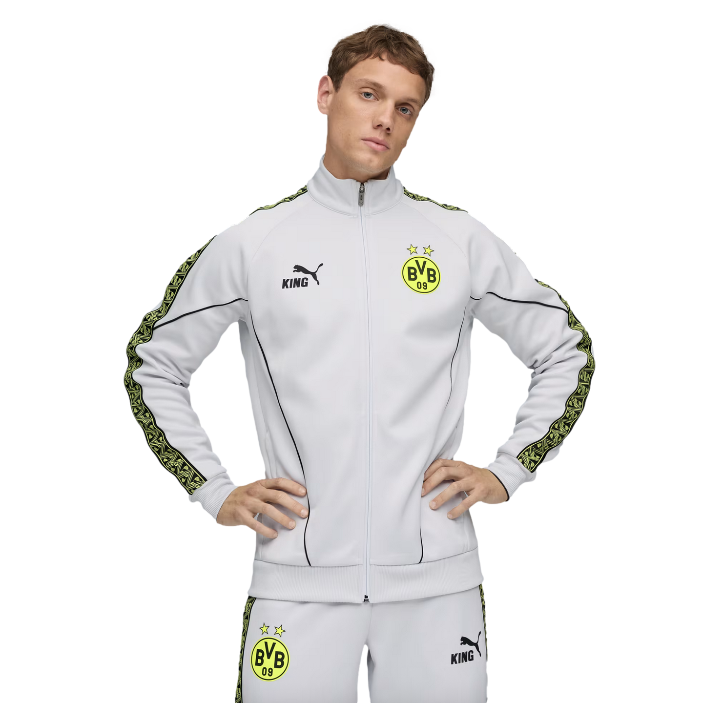 A man in a light grey Puma BVB 09 tracksuit with yellow patterned side stripes, posing against a black background with hands on his hips.