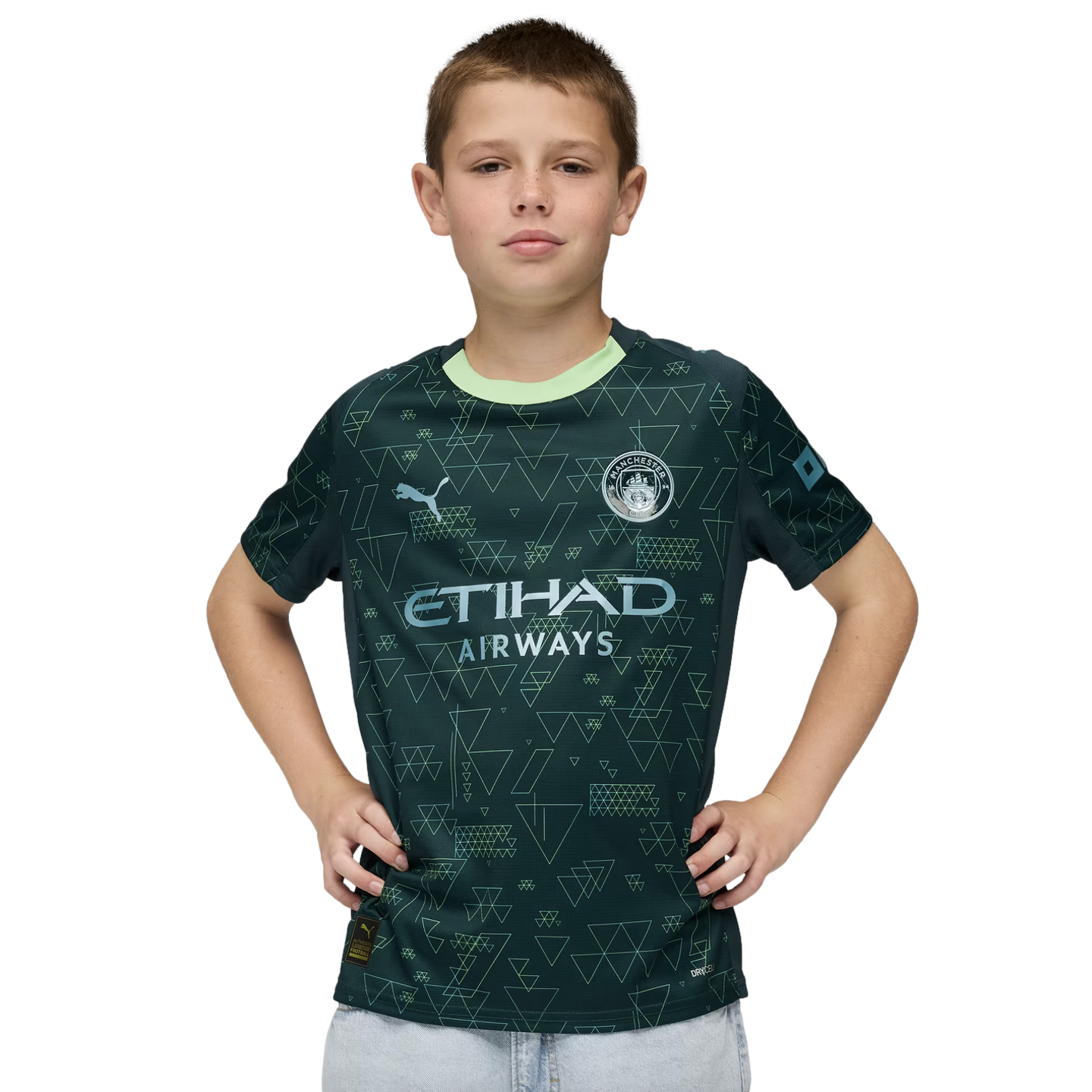 A young boy posing in a dark green Manchester City soccer jersey with geometric triangle patterns, hands on his hips against a black background.