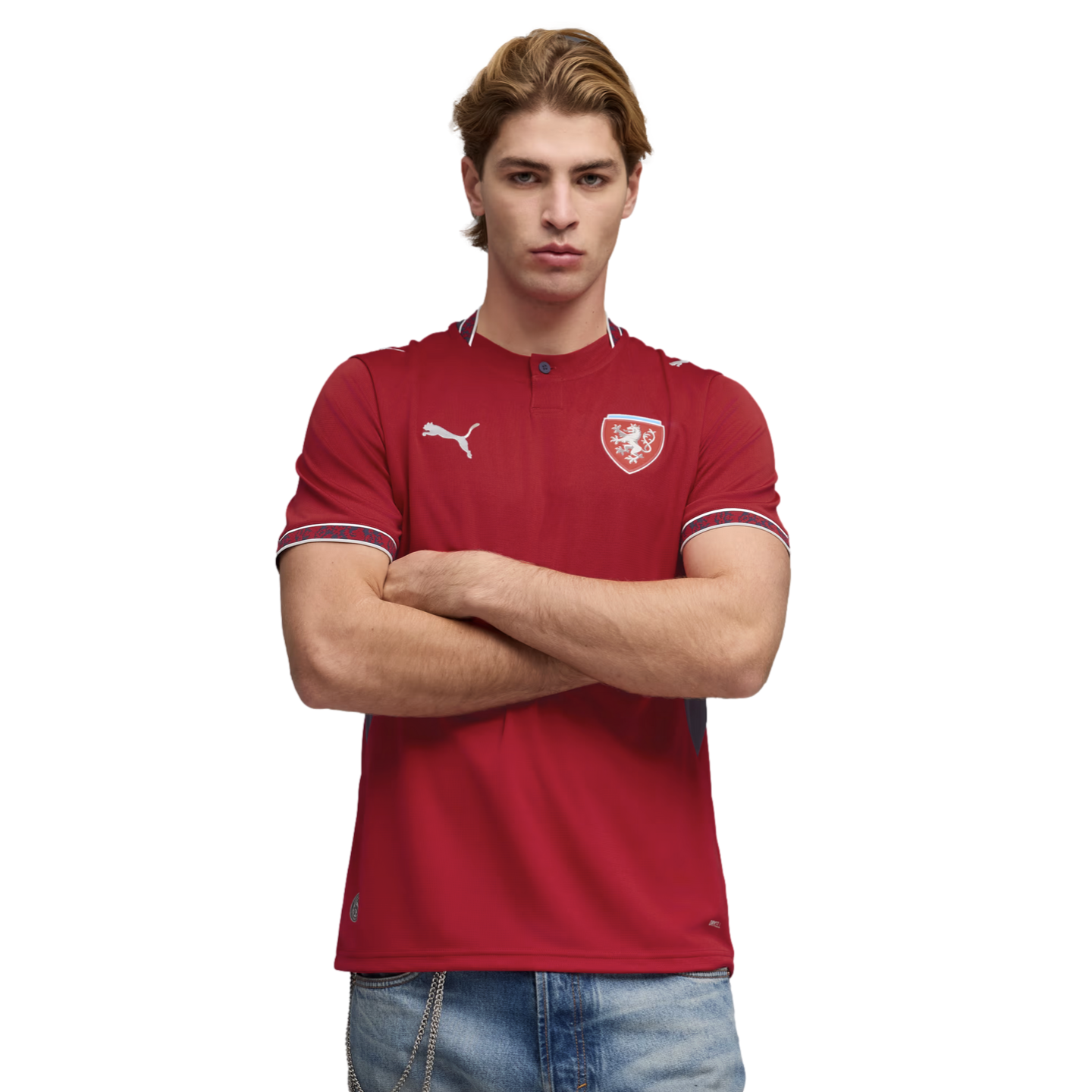 A man with crossed arms wears a red Puma Czech Republic soccer jersey and light-wash jeans against a black background.