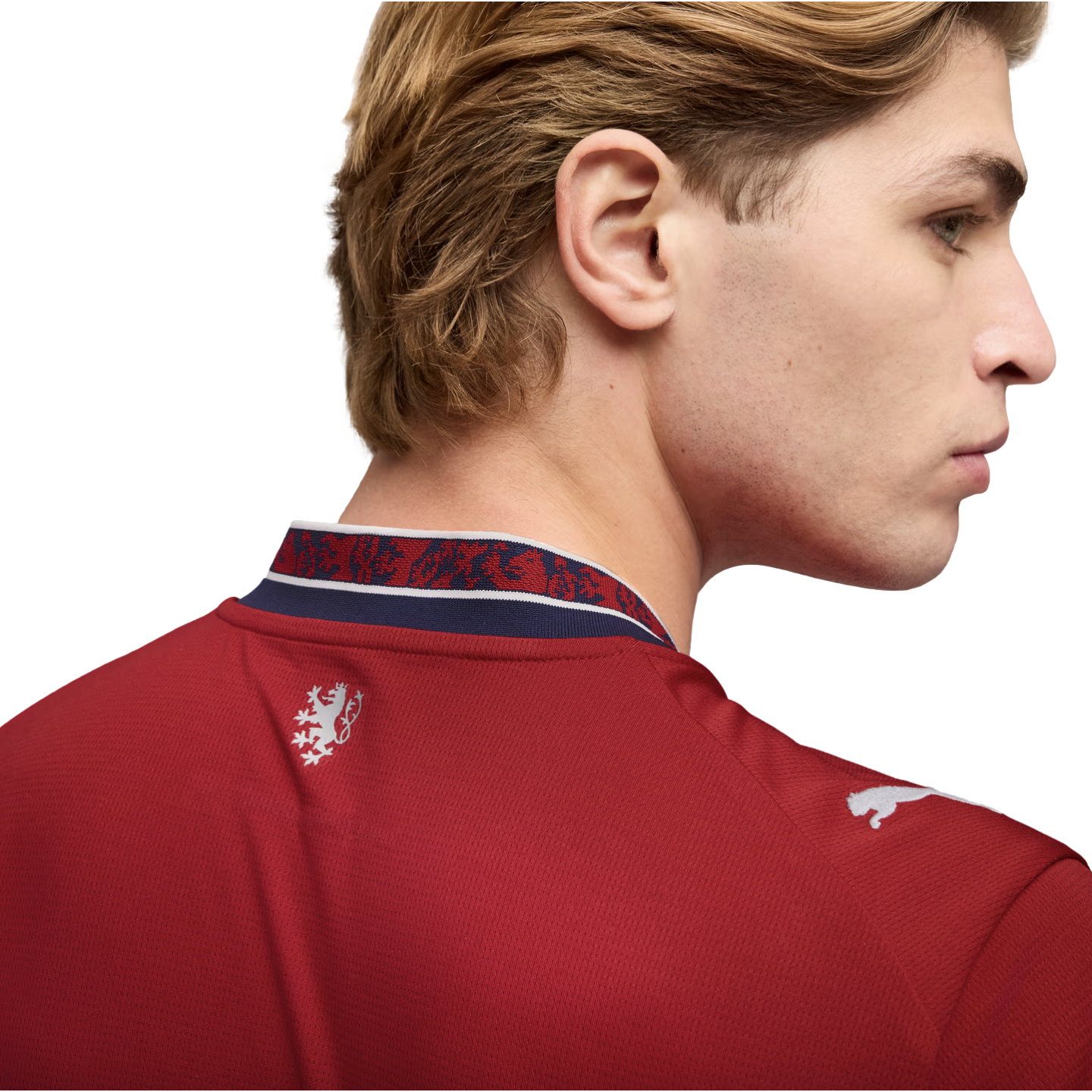 Man with wavy blonde hair in profile wearing a red Puma jersey with a white lion crest and patterned collar against a black background.