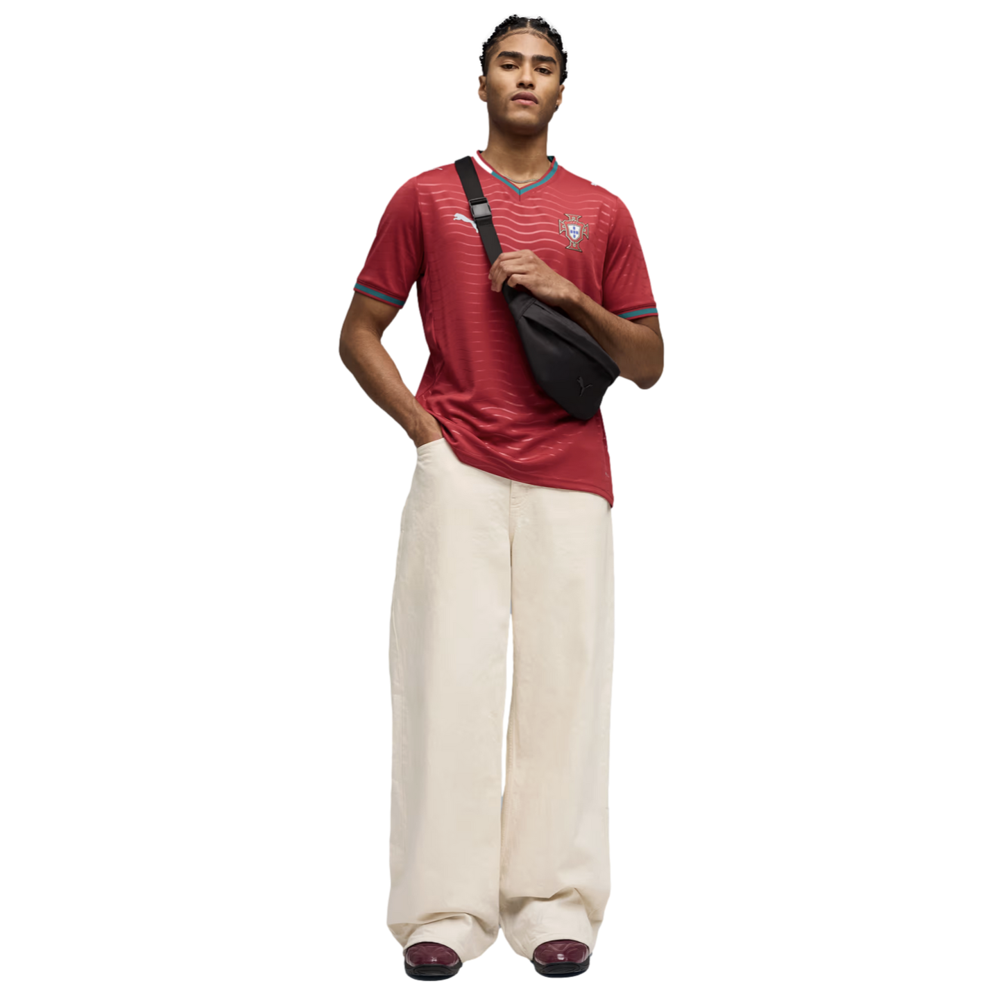 A man wearing a red Portugal football jersey, cream wide-leg trousers, and a black crossbody bag stands against a black background.