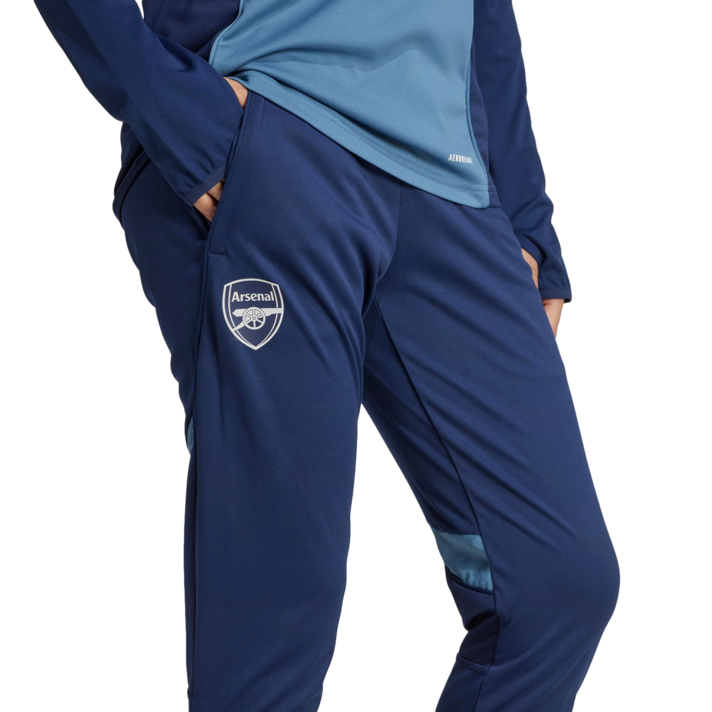 A person wears navy blue Arsenal training pants with the club crest on the right leg, paired with a two-tone blue long-sleeve top. The background is black.