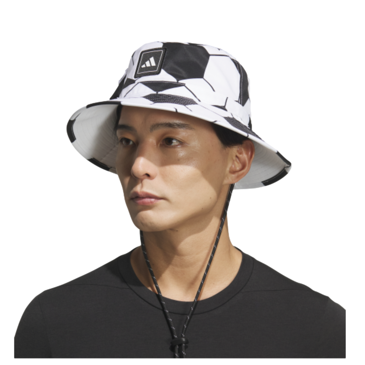A man wears a white Adidas bucket hat with a black soccer ball pattern and a chin strap, set against a black background.