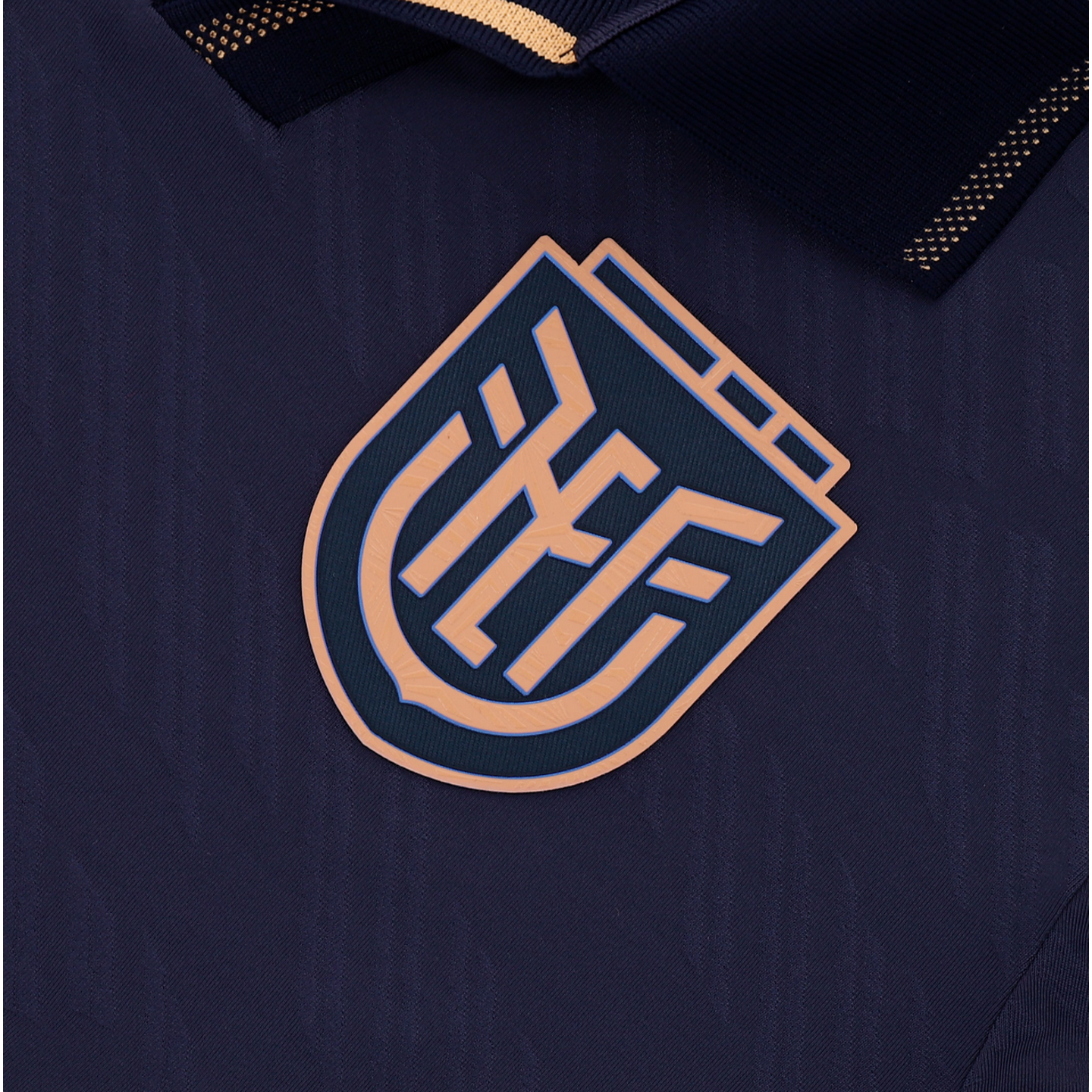 A close-up of a dark navy blue athletic jersey featuring a raised, shield-shaped logo with stylized, interlocking tan letters. The jersey fabric has a subtle wavy texture, and a dark collar with gold accents is partially visible at the top.