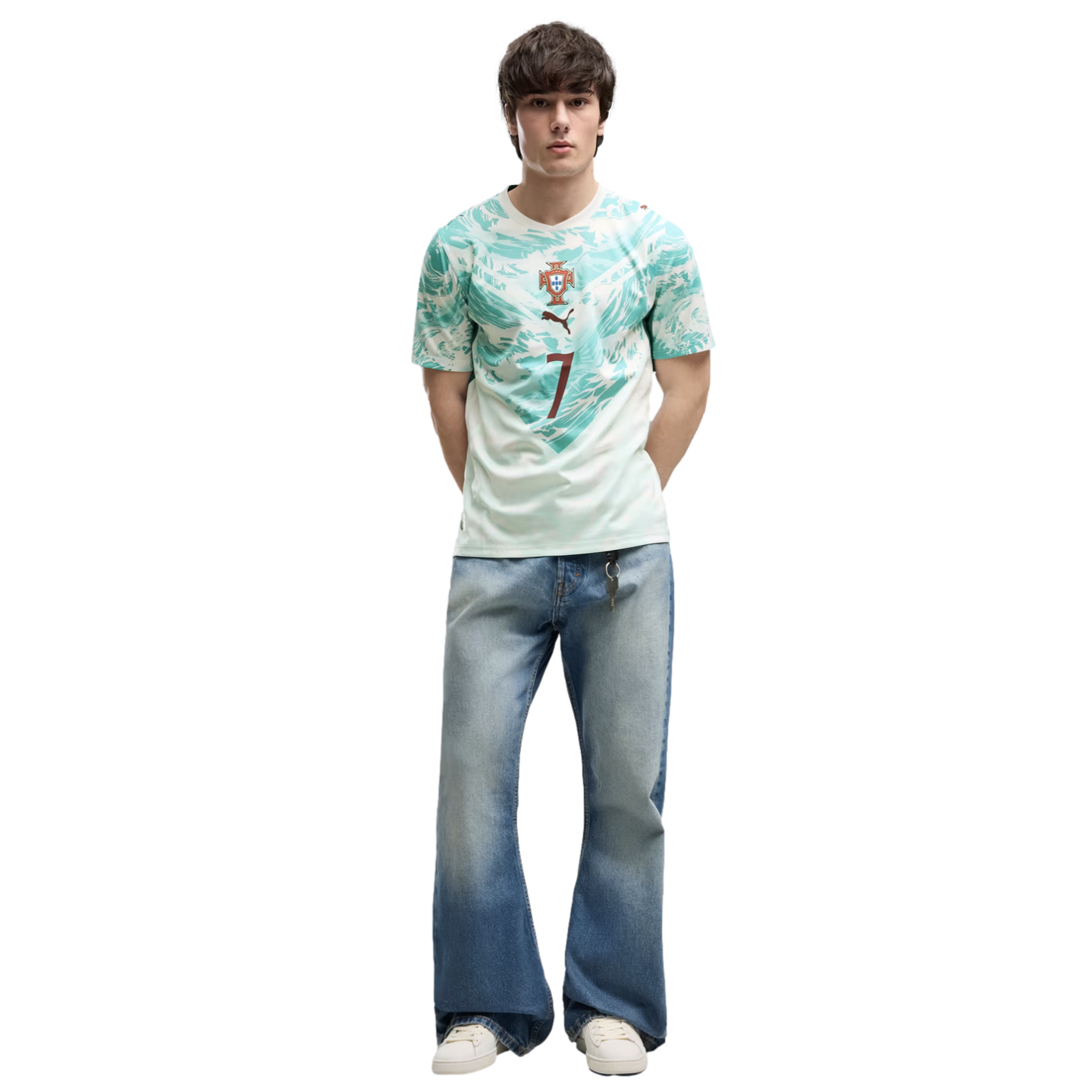 A young man with dark, textured hair stands against a solid black background. He is wearing a light green and teal patterned athletic jersey with a crest and the number 7, wide-leg light-wash blue jeans, and white sneakers. He stands with his hands behind
