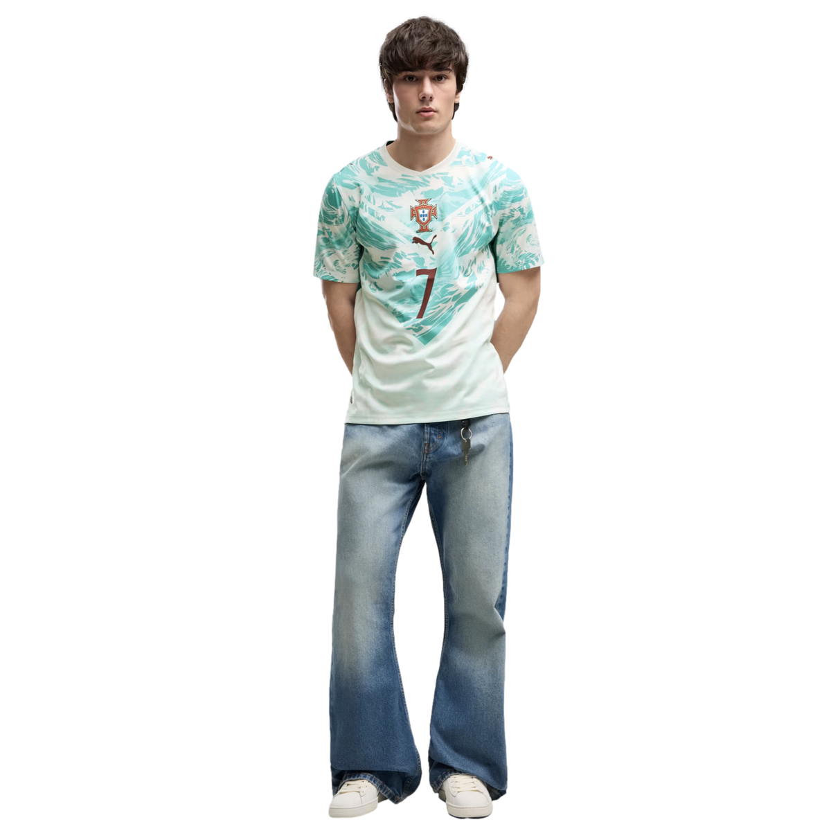 A young man with dark, textured hair stands against a solid black background. He is wearing a light green and teal patterned athletic jersey with a crest and the number 7, wide-leg light-wash blue jeans, and white sneakers. He stands with his hands behind