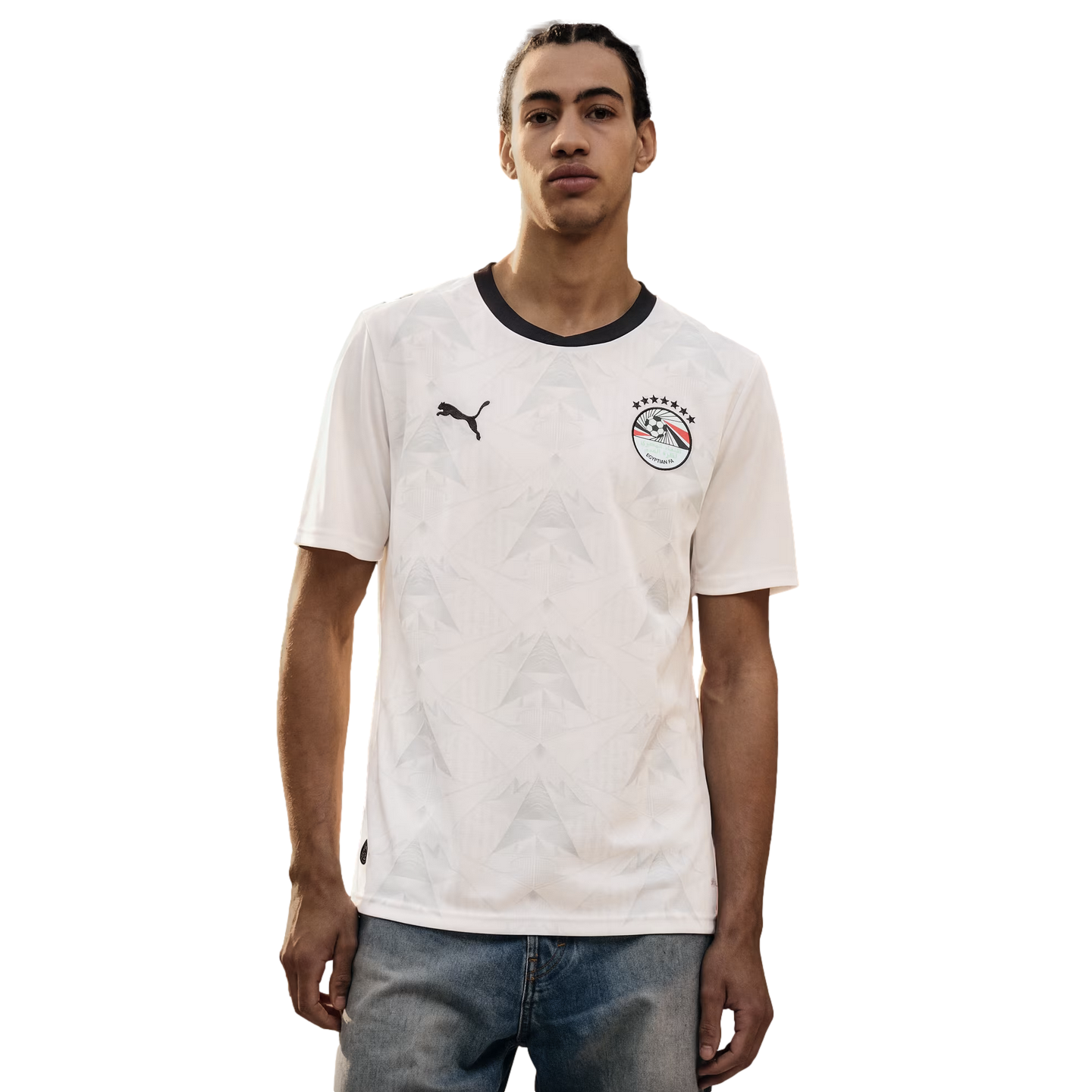 A young man wearing a white Puma Egypt national football team away jersey with a black collar and a tonal geometric pattern, paired with blue jeans against a solid black background.
