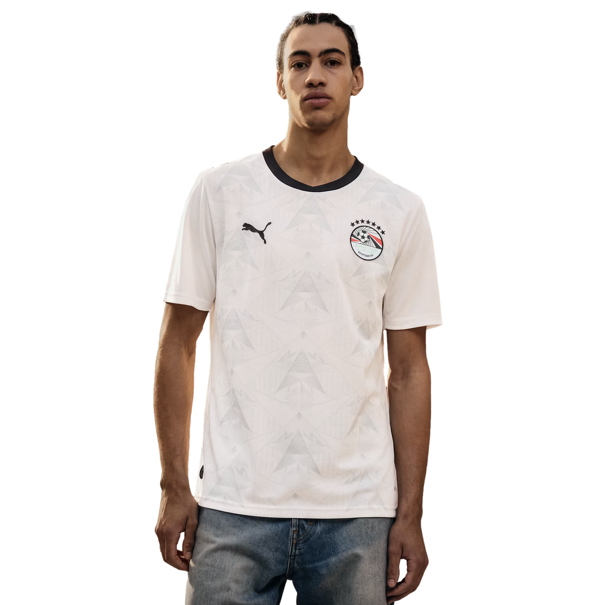 A young man wearing a white Puma Egypt national football team away jersey with a black collar and a tonal geometric pattern, paired with blue jeans against a solid black background.