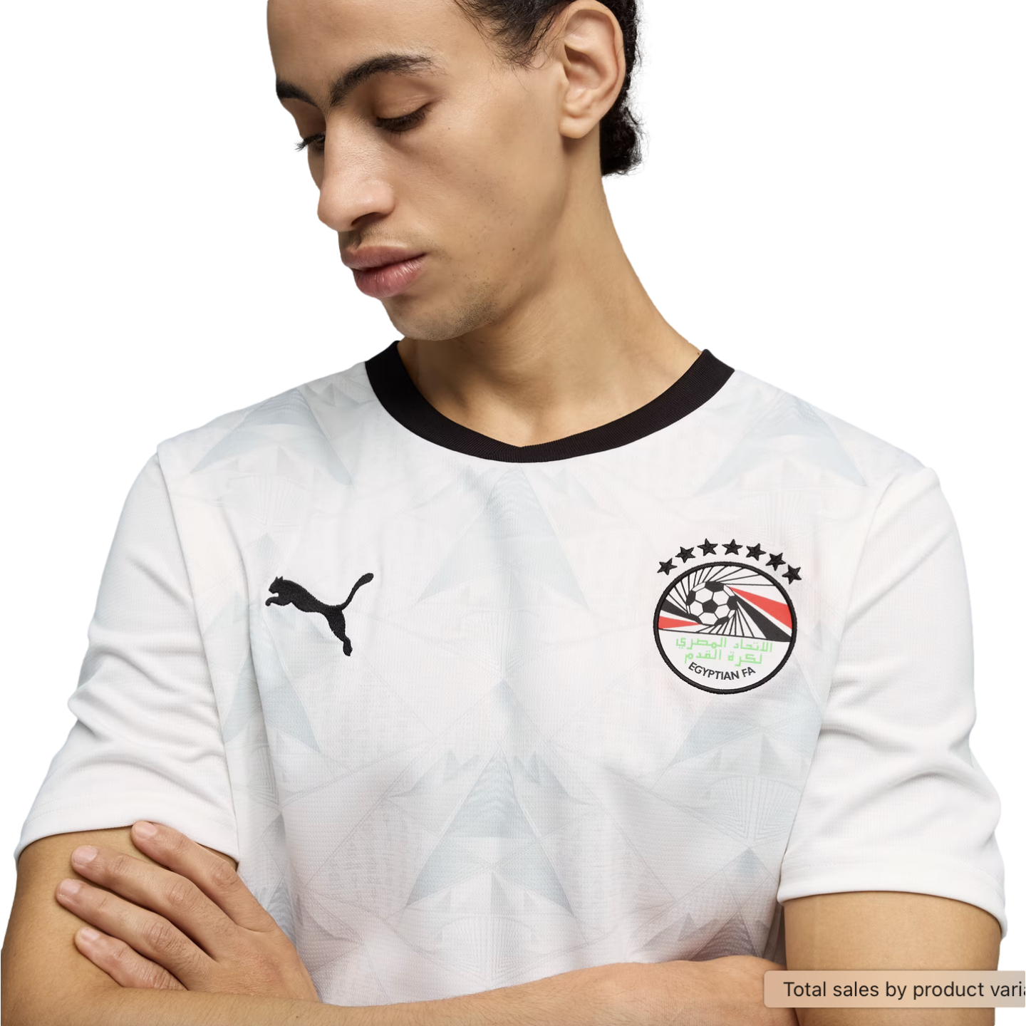 A young man looks down with his arms crossed, wearing a white Puma soccer jersey with a black crew neck and a subtle geometric pattern. The jersey features the Egyptian Football Association crest on the left chest and a black Puma logo on the right, all s