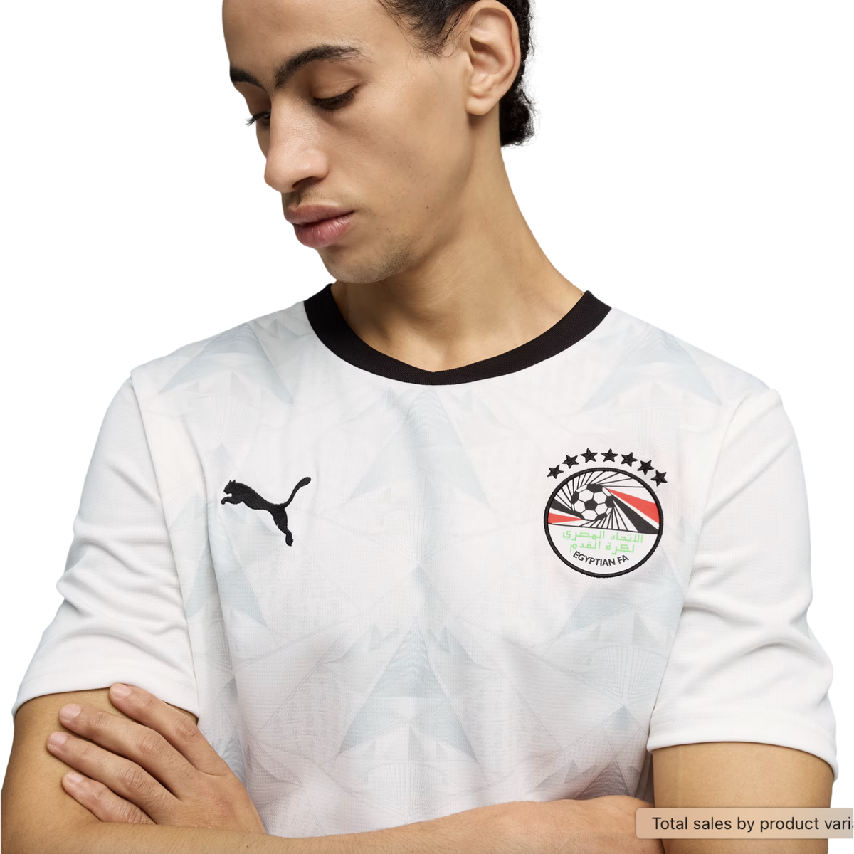 A young man looks down with his arms crossed, wearing a white Puma soccer jersey with a black crew neck and a subtle geometric pattern. The jersey features the Egyptian Football Association crest on the left chest and a black Puma logo on the right, all s