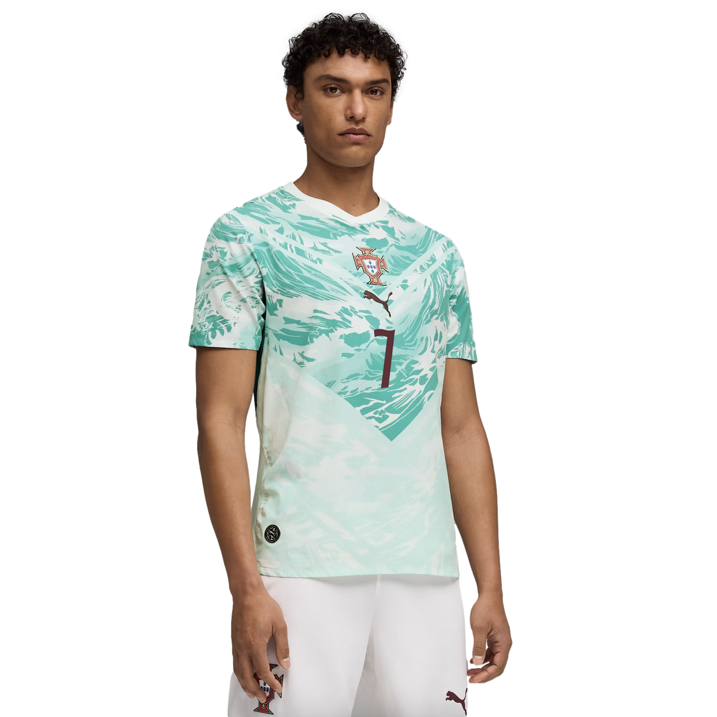 A young man models a white and light green abstract-patterned soccer jersey with a central crest and the number 7, paired with white shorts, against a black background.