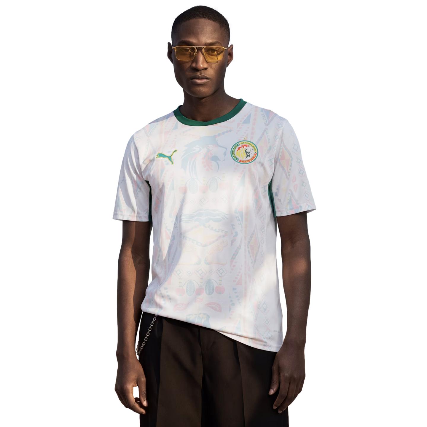 A man wearing yellow-tinted sunglasses and a white Senegal national team football jersey with a green collar, standing against a solid black background.