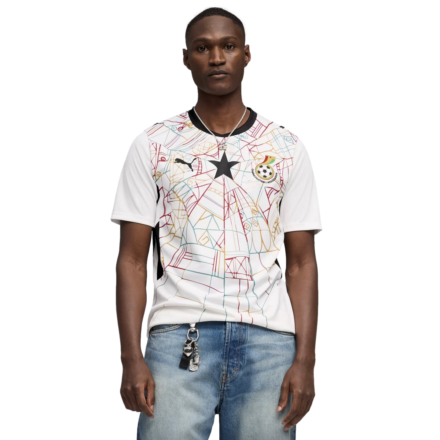 A medium shot of a Black man wearing a white Puma Ghana national football team jersey with a colorful geometric pattern and a central black star. He is also wearing light-wash blue jeans and a silver necklace, set against a solid black background.