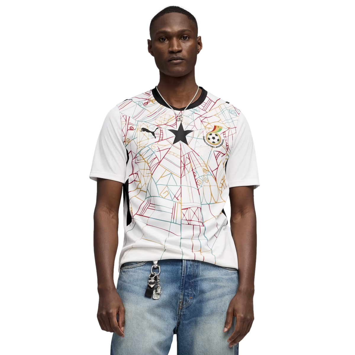 A medium shot of a Black man wearing a white Puma Ghana national football team jersey with a colorful geometric pattern and a central black star. He is also wearing light-wash blue jeans and a silver necklace, set against a solid black background.