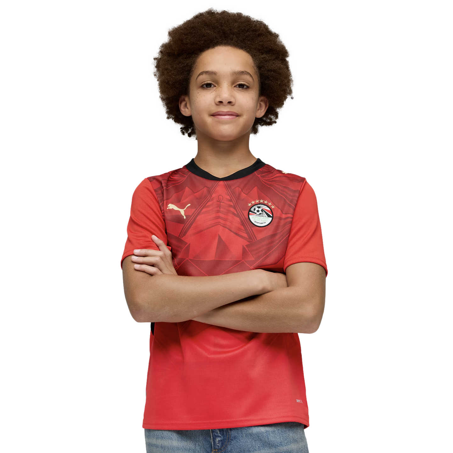 A young person with a large afro and a slight smile poses with their arms crossed. They are wearing a red Egyptian national team soccer jersey and blue jeans against a solid black background.