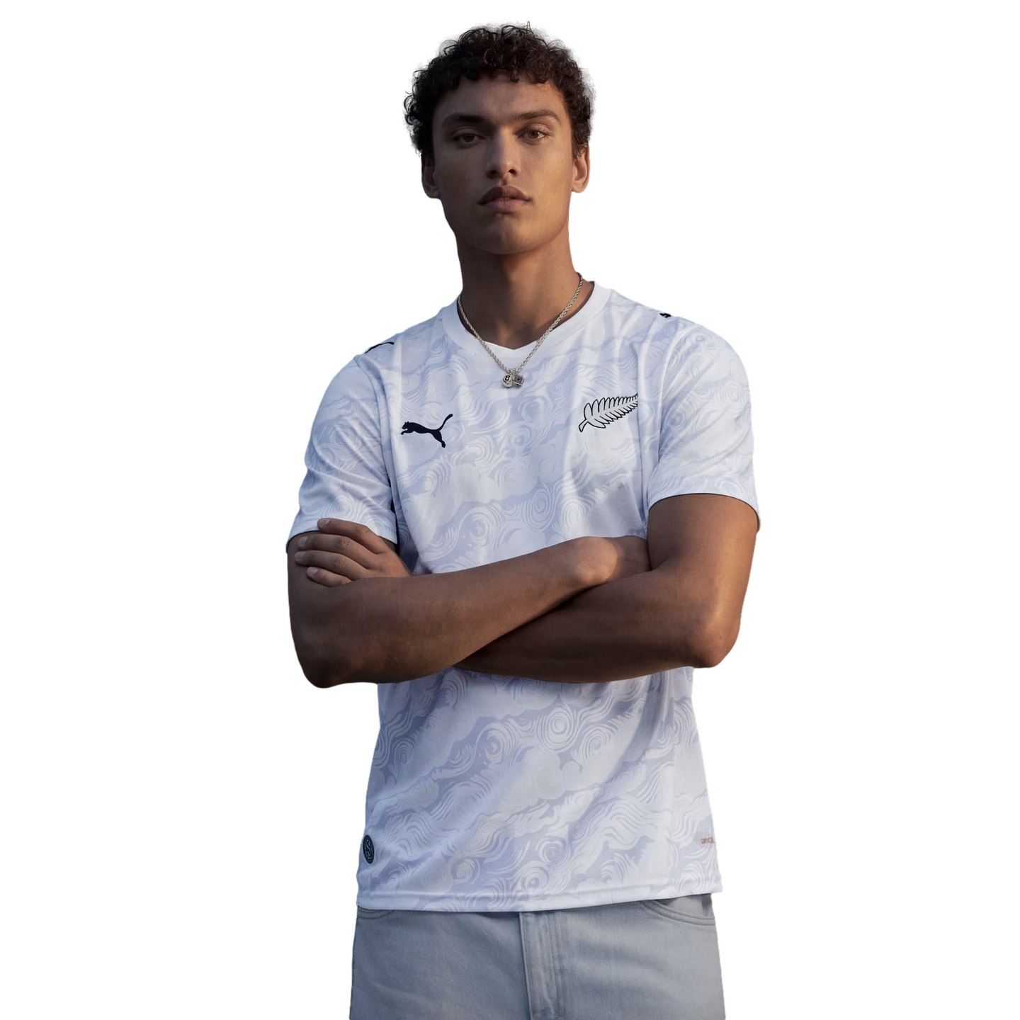 A waist-up portrait of a young man with curly hair posing with his arms crossed against a solid black background. He is wearing a white Puma jersey with a subtle, light-grey tonal swirl pattern. The jersey features a black Puma logo on the right chest and