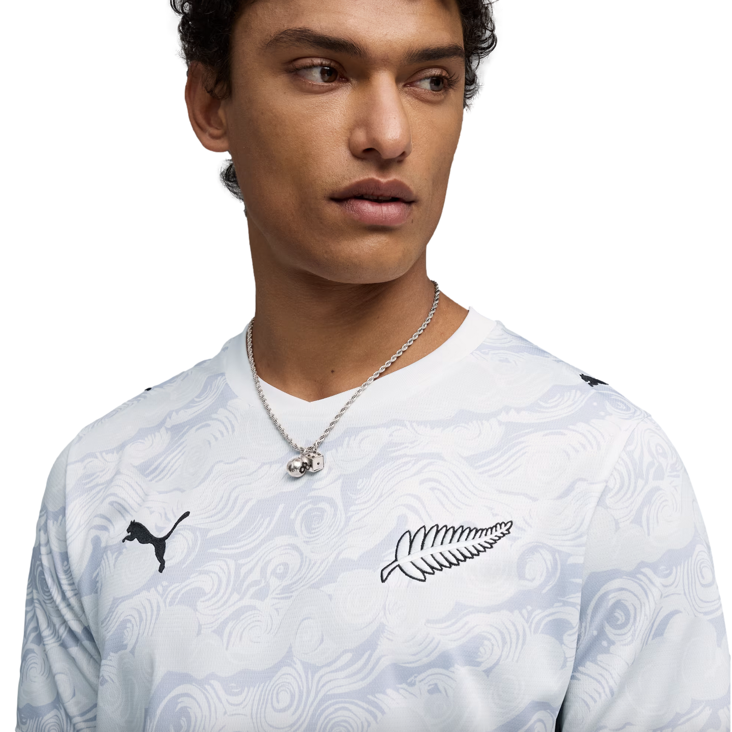 A close-up of a young man wearing a white, wavy-patterned long-sleeve Puma shirt. The shirt features a black Puma logo on the right side and a black silver fern logo on the left. He is also wearing a silver chain necklace with a dice pendant against a sol
