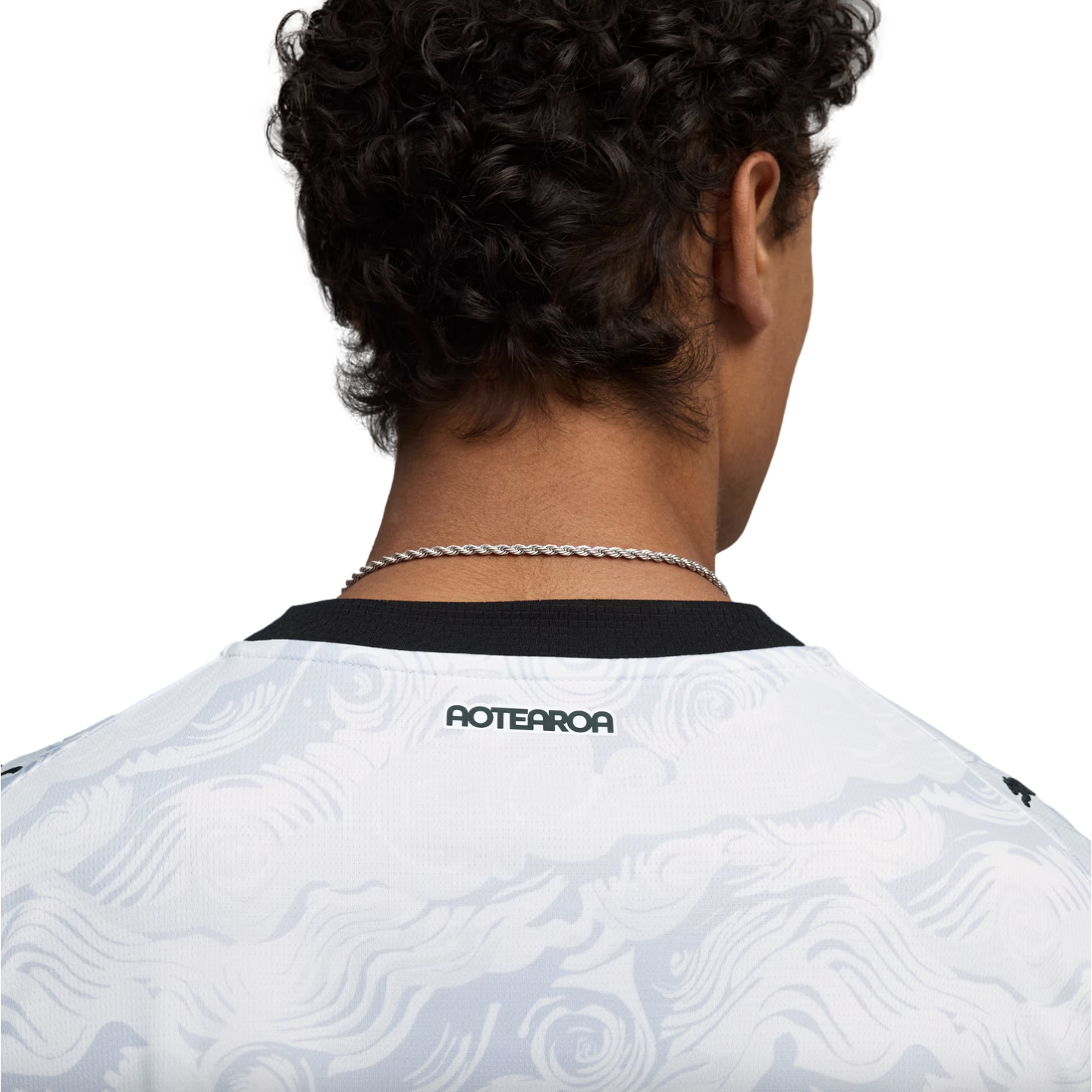 Close-up from behind of a person with dark, curly hair wearing a white jersey with a grey swirl pattern and a black collar. The word "AOTEAROA" is printed in black on the back of the jersey, just below the collar. A thin silver chain necklace is visible a