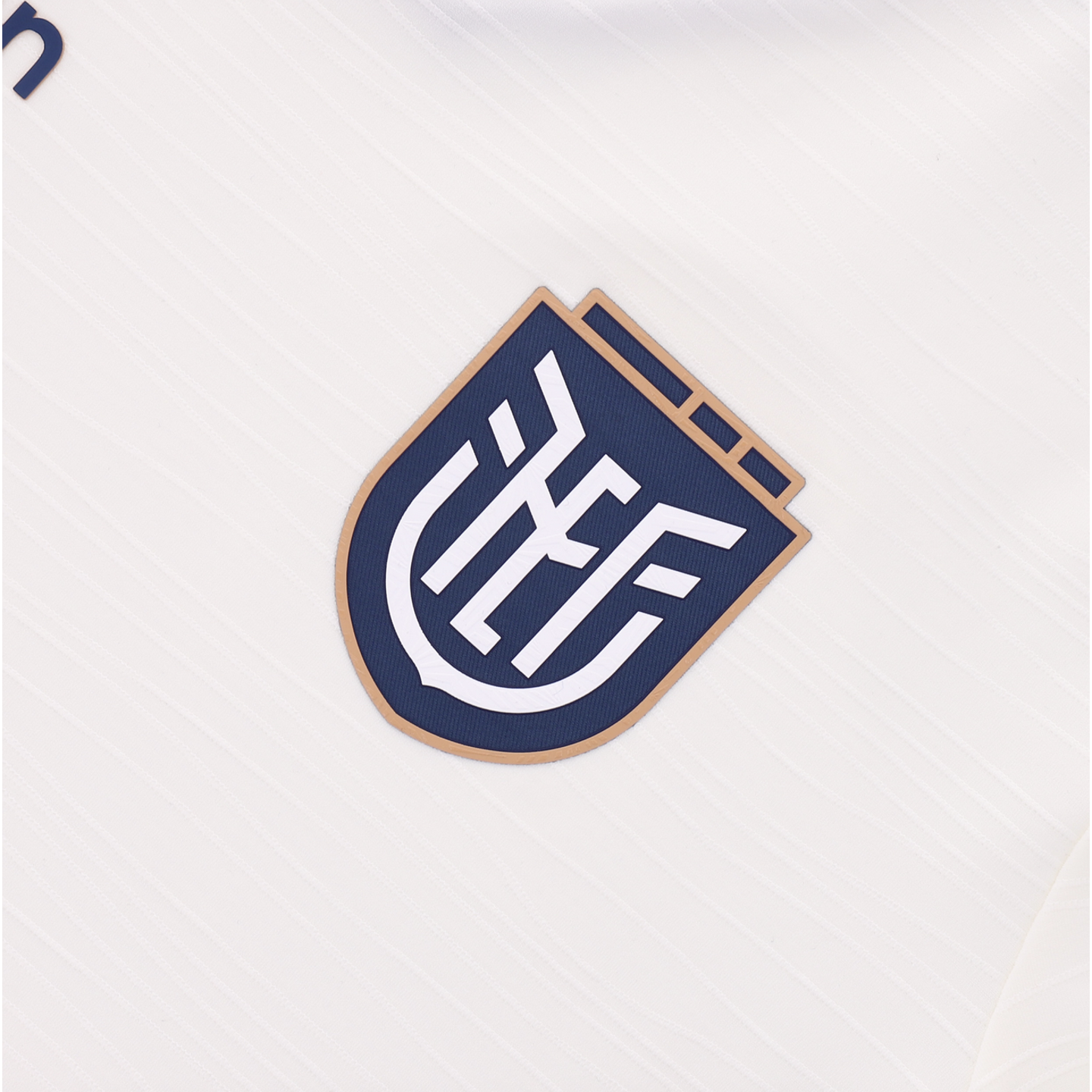 A close-up of the navy blue and tan FEF (Federación Ecuatoriana de Fútbol) crest on a white textured jersey.