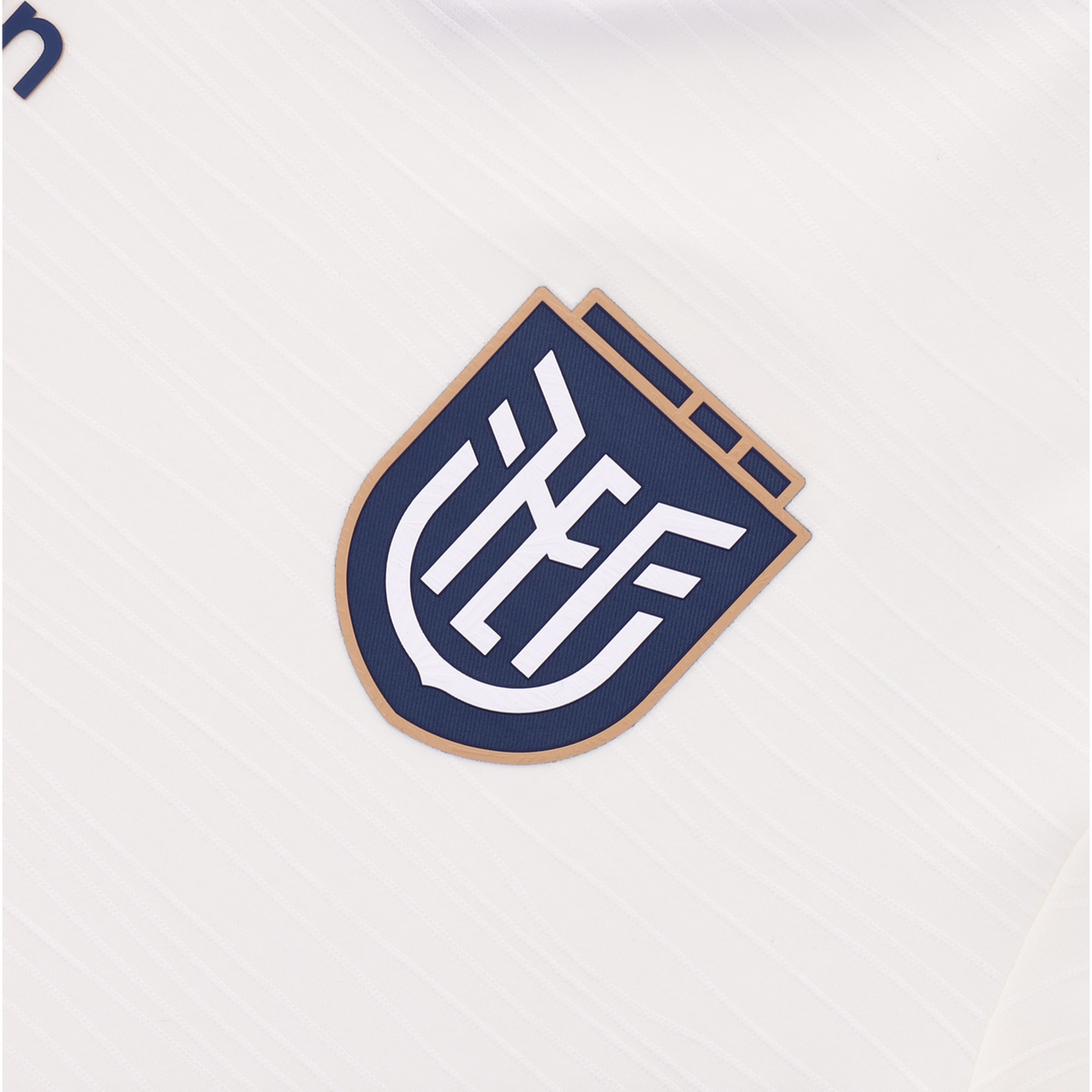 A close-up of the navy blue and tan FEF (Federación Ecuatoriana de Fútbol) crest on a white textured jersey.