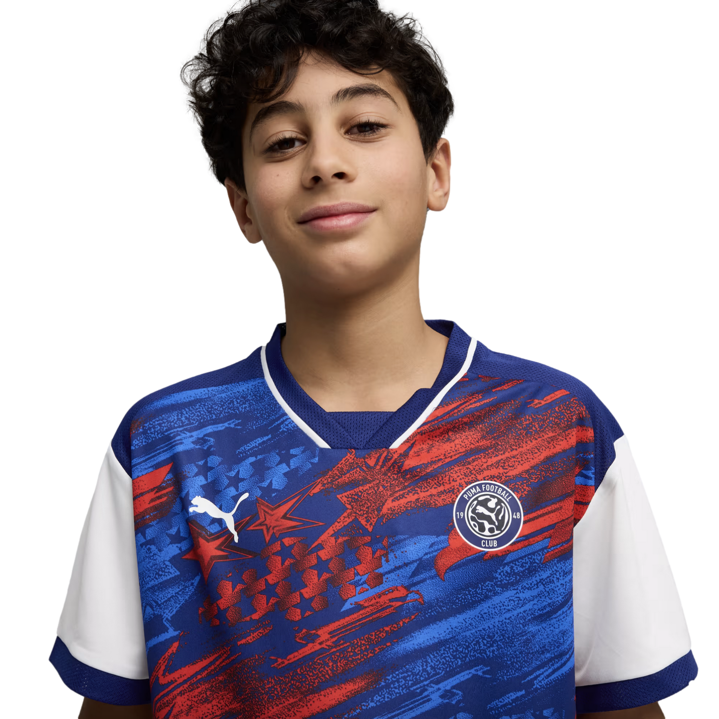 A young boy with dark curly hair and a slight smile, wearing a blue and red patterned Puma Football Club soccer jersey against a solid black background.