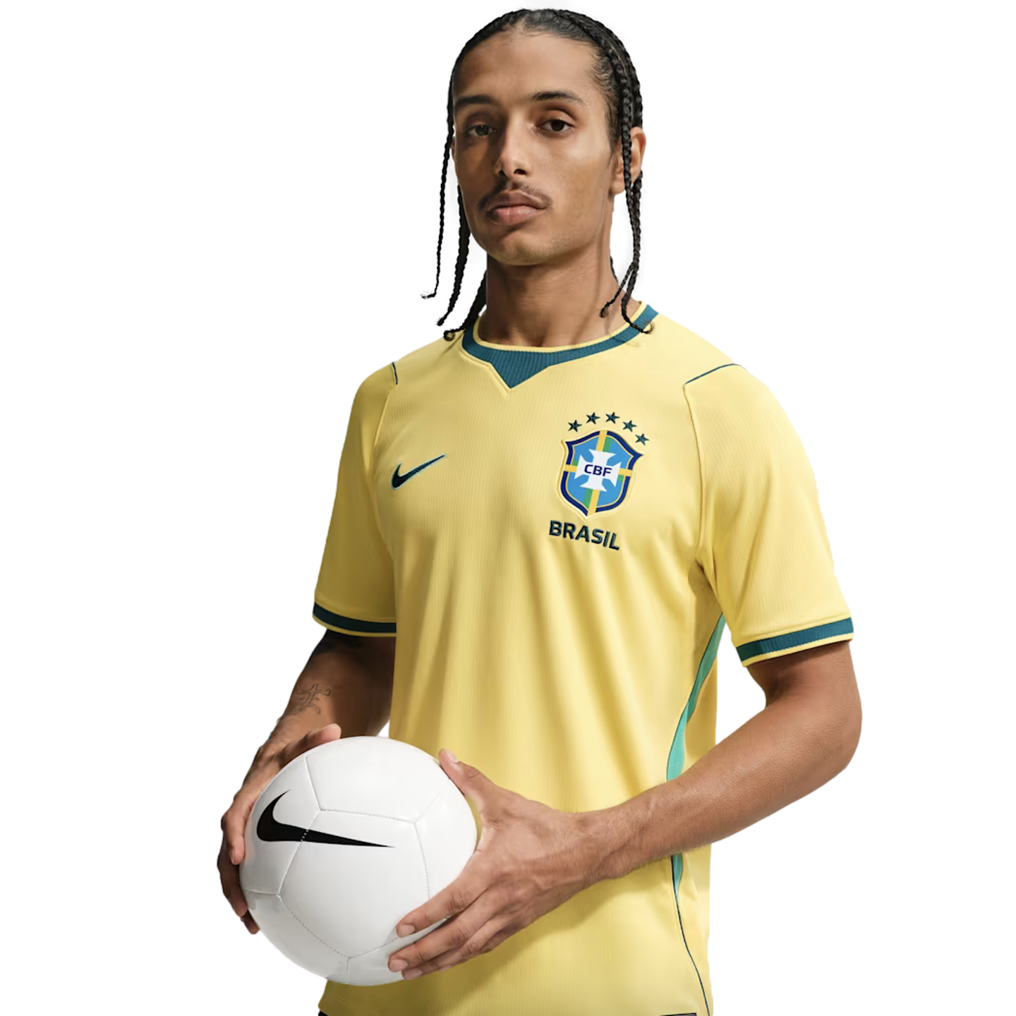 A man with braided hair wearing a yellow Nike Brazil national football team jersey, holding a white soccer ball against a solid black background.