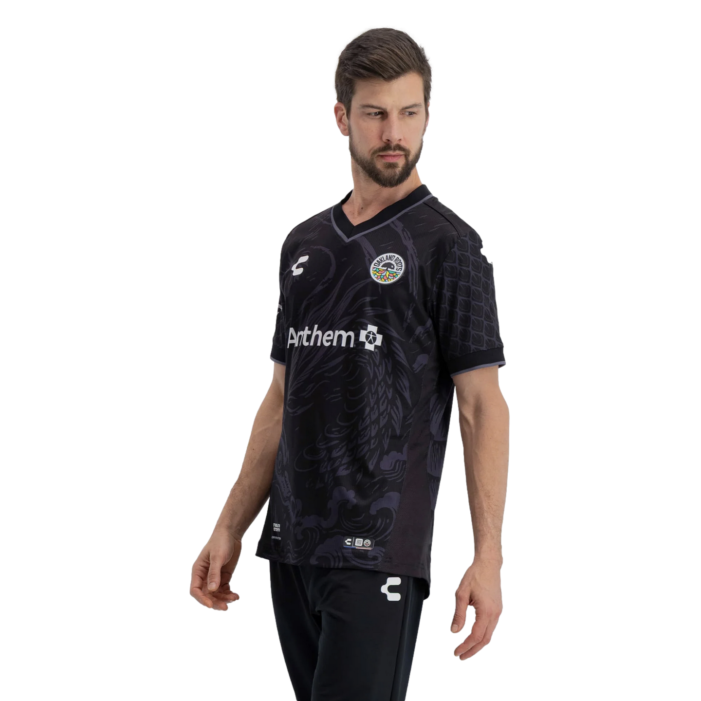 A man models an Oakland Roots SC soccer jersey against a solid black background. The black V-neck jersey features tonal grey wavy patterns on the front, textured scale-like patterns on the sleeves, the team's circular crest on the chest, and an Anthem spo