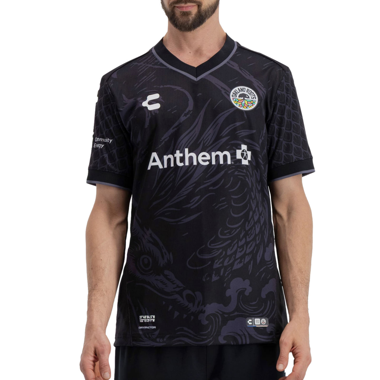 A man wearing a black Oakland Roots soccer jersey featuring a subtle tonal dragon graphic. The jersey has a white "Anthem" sponsor logo across the center, the team crest on the left chest, and geographic coordinates printed at the bottom left.
