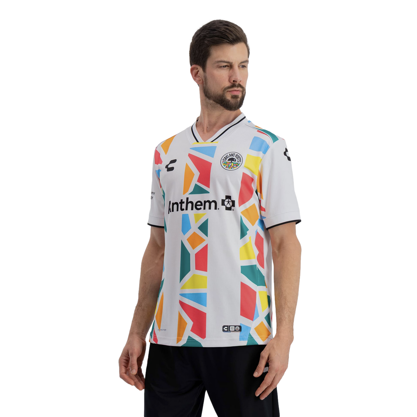 A man models a white Oakland Roots soccer jersey featuring a vibrant, multi-colored geometric mosaic pattern in shades of red, yellow, orange, blue, and green. The word "Anthem" is printed in black across the chest, and the jersey includes a black-and-whi