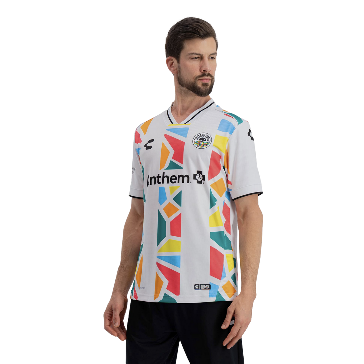 A man models a white Oakland Roots soccer jersey featuring a vibrant, multi-colored geometric mosaic pattern in shades of red, yellow, orange, blue, and green. The word "Anthem" is printed in black across the chest, and the jersey includes a black-and-whi