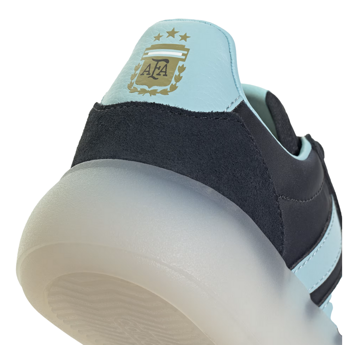 A close-up shot focusing on the heel of an Adidas shoe, featuring a light blue panel with the gold Argentina Football Association (AFA) crest and three stars. Below the blue panel is a dark navy textured material and a thick, semi-translucent white sole,