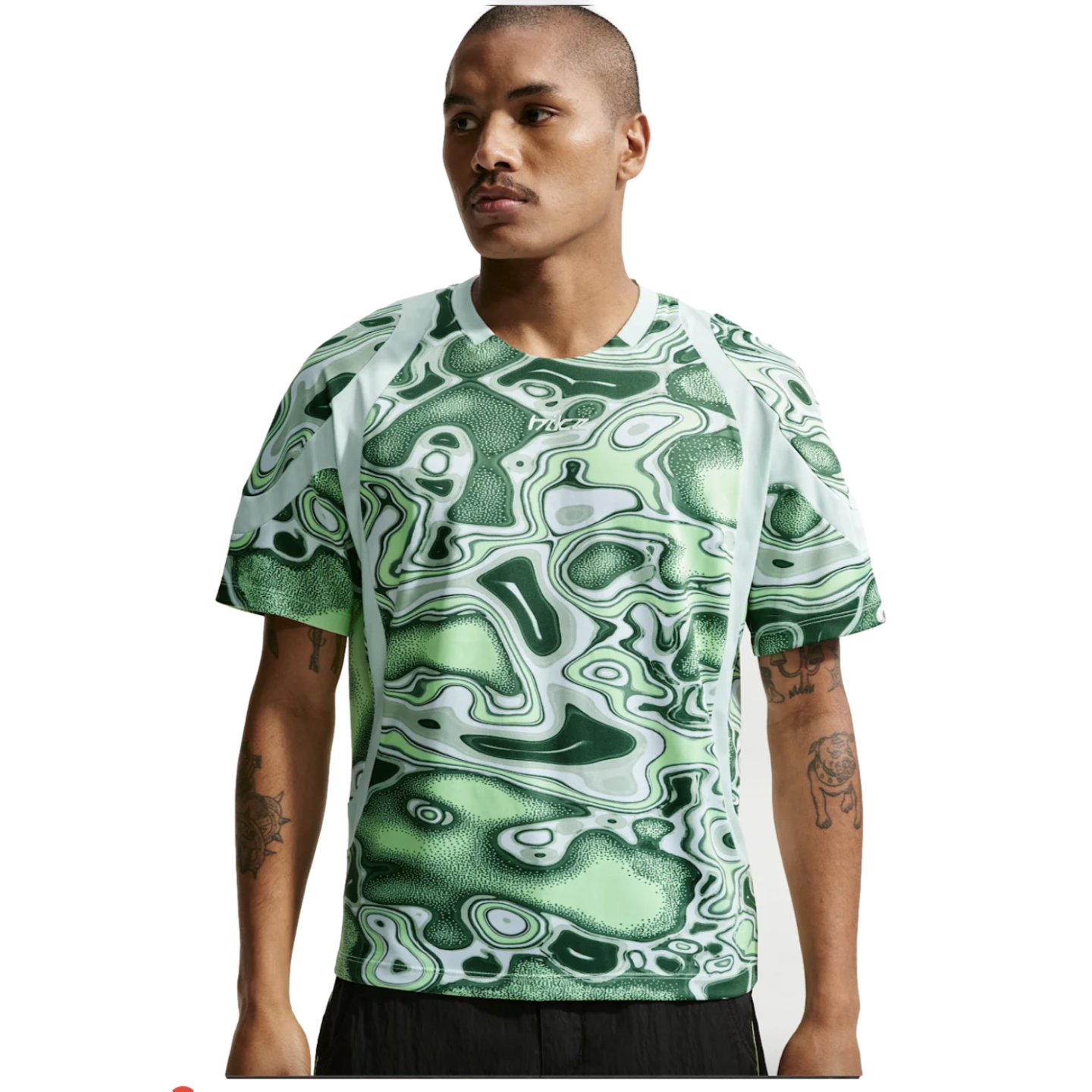 A man wearing a green and white abstract-patterned t-shirt with raglan sleeves and black shorts against a white background.