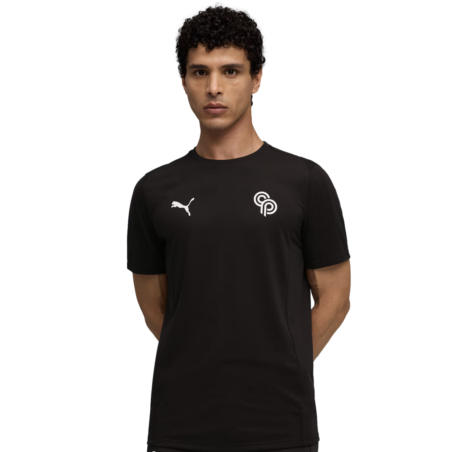 Man with curly dark hair in a black Puma tee with white logos on the chest, posing with hands behind his back against a plain black background.