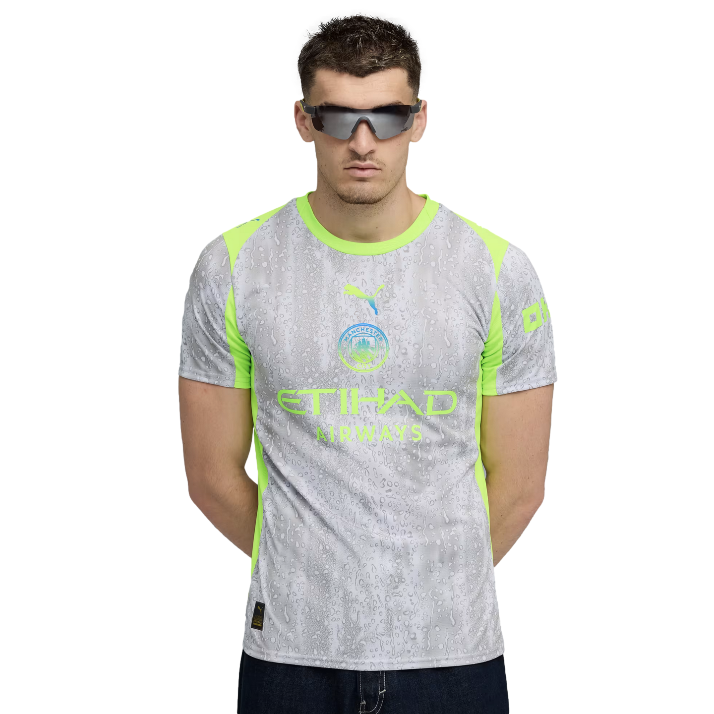 A man in dark sunglasses stands against a black background wearing a grey and lime green Manchester City jersey with a water droplet pattern.