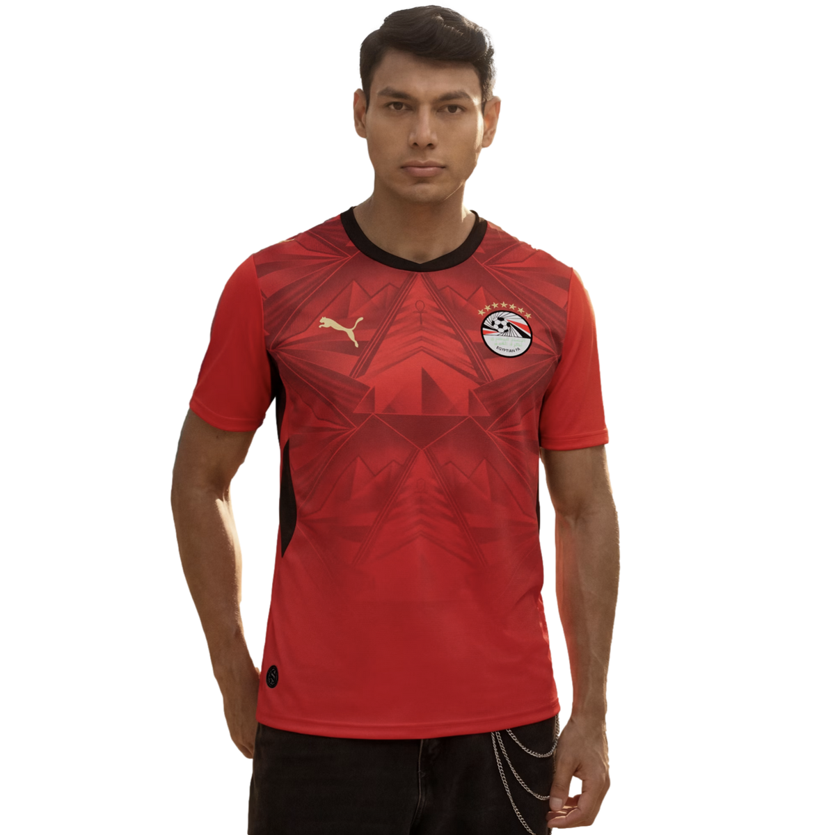 A man wearing a red Egypt national football team home jersey by Puma, featuring subtle geometric patterns and the team crest, standing against a plain black background.