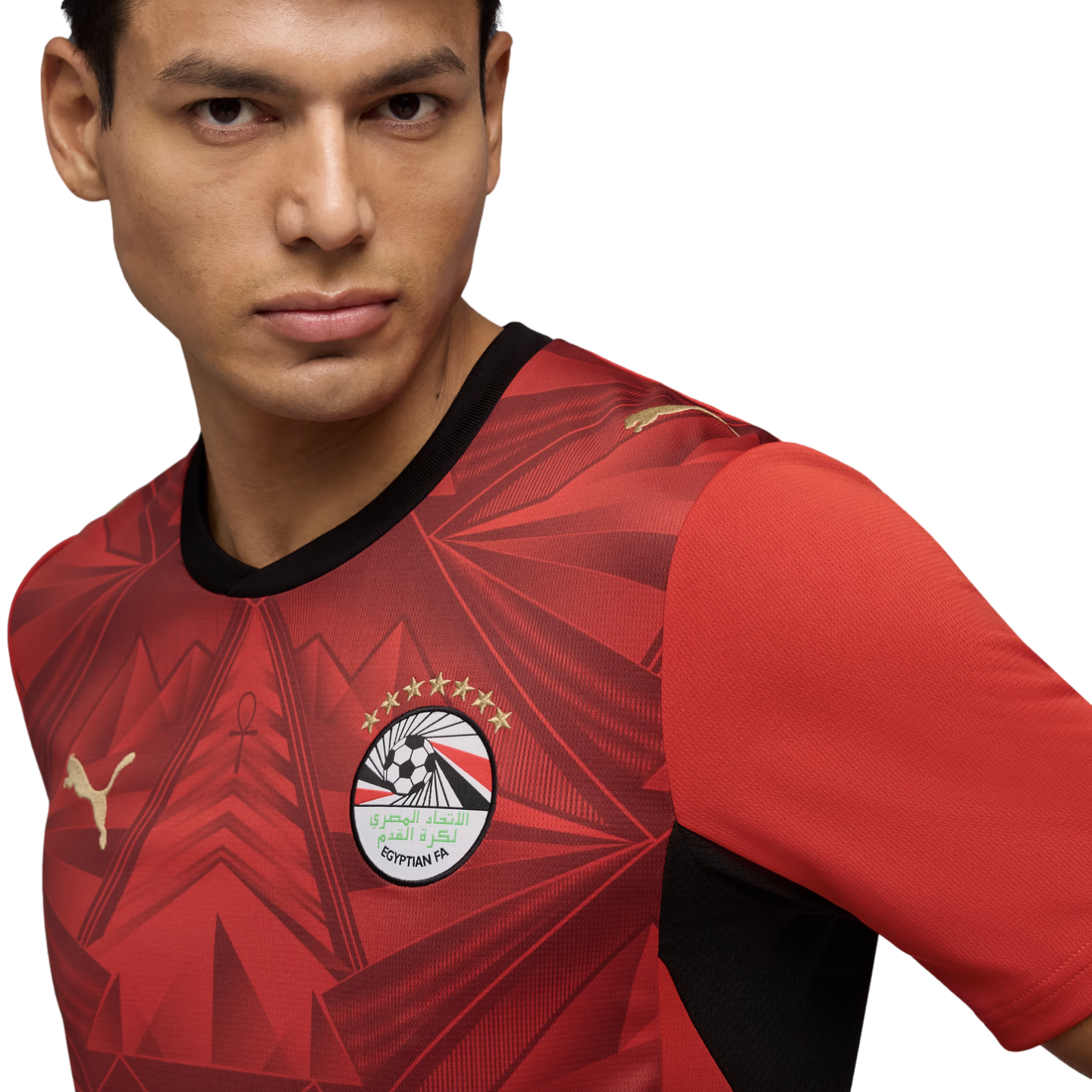 A close-up portrait of a man wearing a red Egypt national football team jersey by Puma. The jersey features an intricate geometric pattern, the Egyptian FA crest with seven gold stars, and gold Puma logos. He looks slightly off-camera against a solid blac