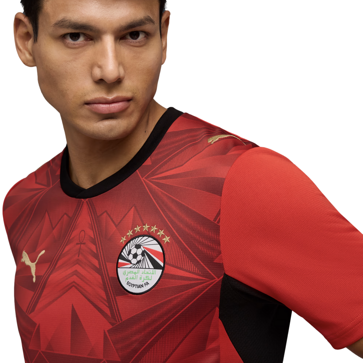 A close-up portrait of a man wearing a red Egypt national football team jersey by Puma. The jersey features an intricate geometric pattern, the Egyptian FA crest with seven gold stars, and gold Puma logos. He looks slightly off-camera against a solid blac