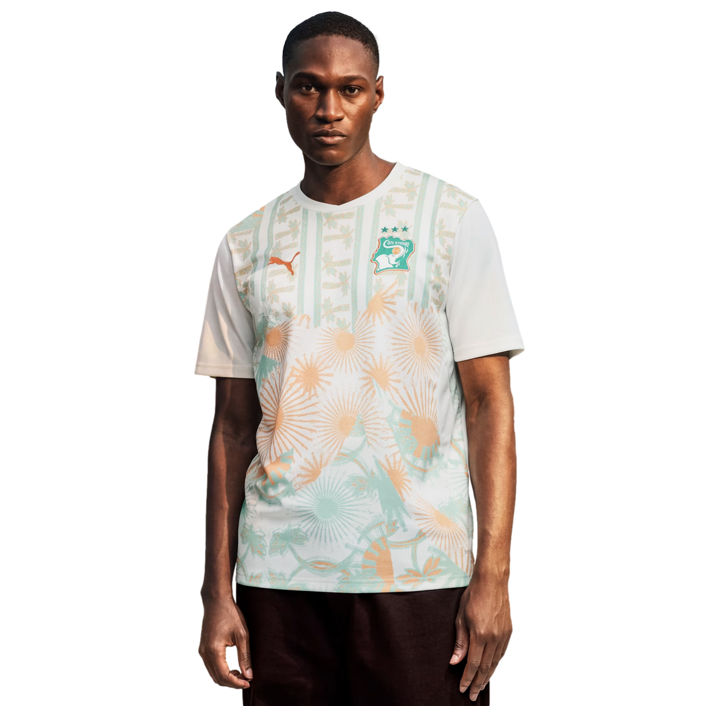 A Black man stands against a plain black background, wearing a white Côte d’Ivoire national football team jersey by Puma. The jersey features an orange Puma logo on the right chest and the team crest on the left, with a light green and orange graphic patt