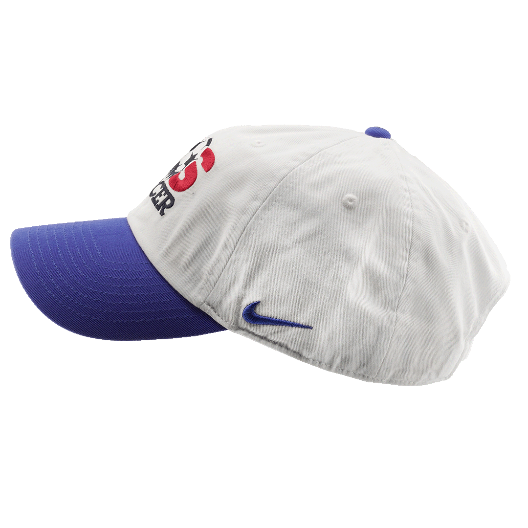 A side view of a white Nike baseball cap with a blue brim and button, featuring a soccer logo on the front and a blue swoosh on the side.