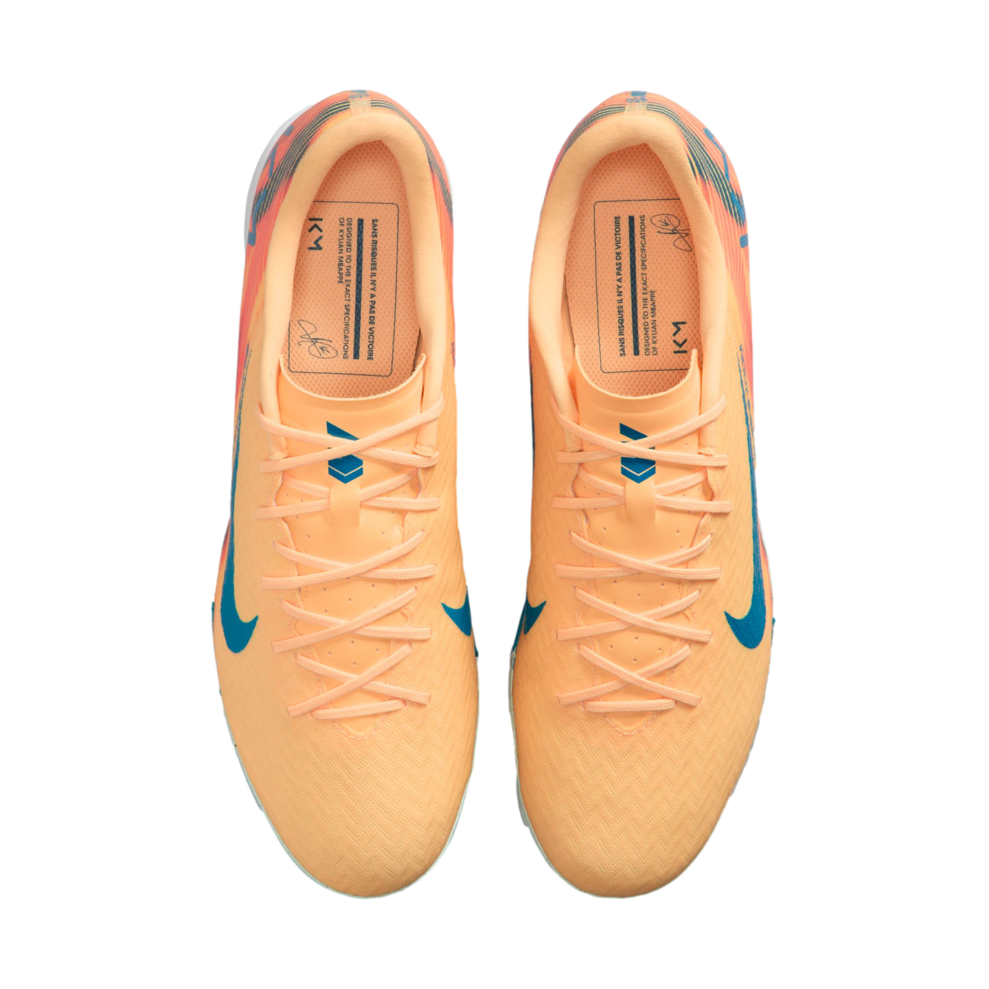 Top-down view of peach-colored Nike soccer cleats with blue accents and "KM" branding, isolated on a black background.