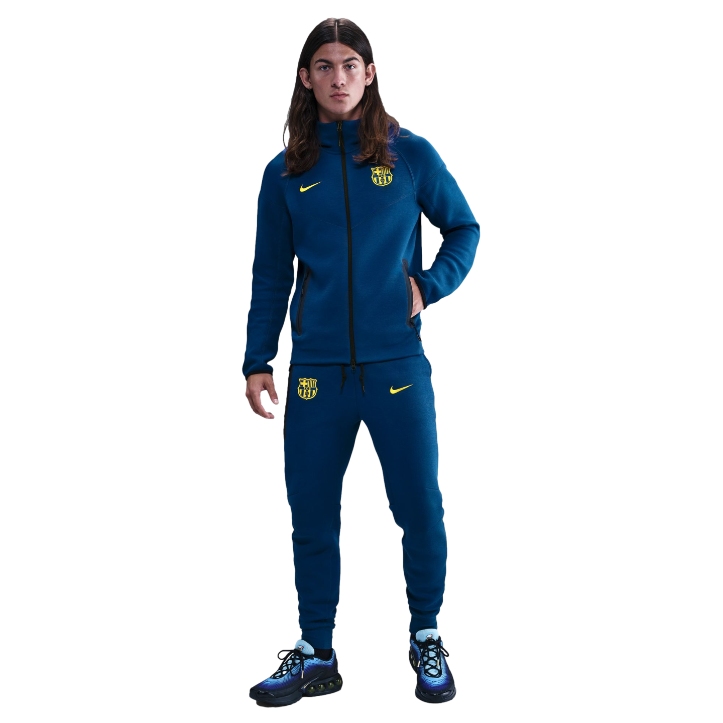 A young man with long hair stands in a blue FC Barcelona Nike tracksuit with yellow logos and blue sneakers against a black background.