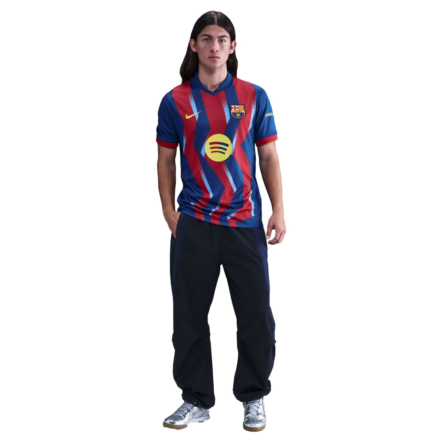 A young man with long hair wearing a red and blue zig-zag FC Barcelona jersey, dark track pants, and silver sneakers against a black background.